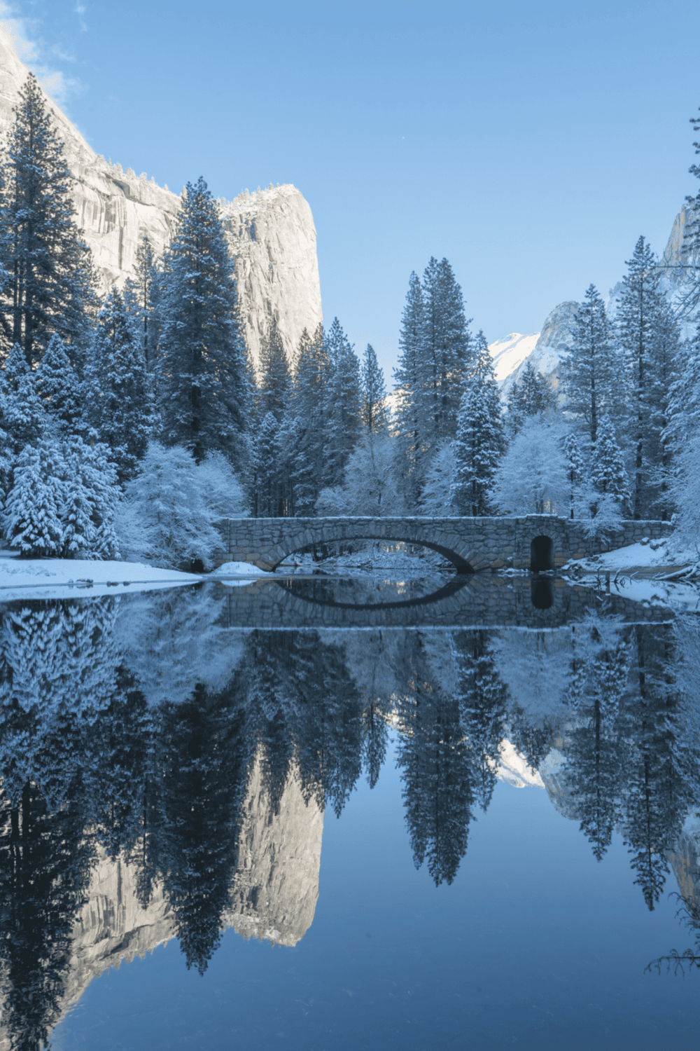 Serene winter landscape with snow-covered trees, mountain cliffs, and a reflective river scene at Yosemite National Park.