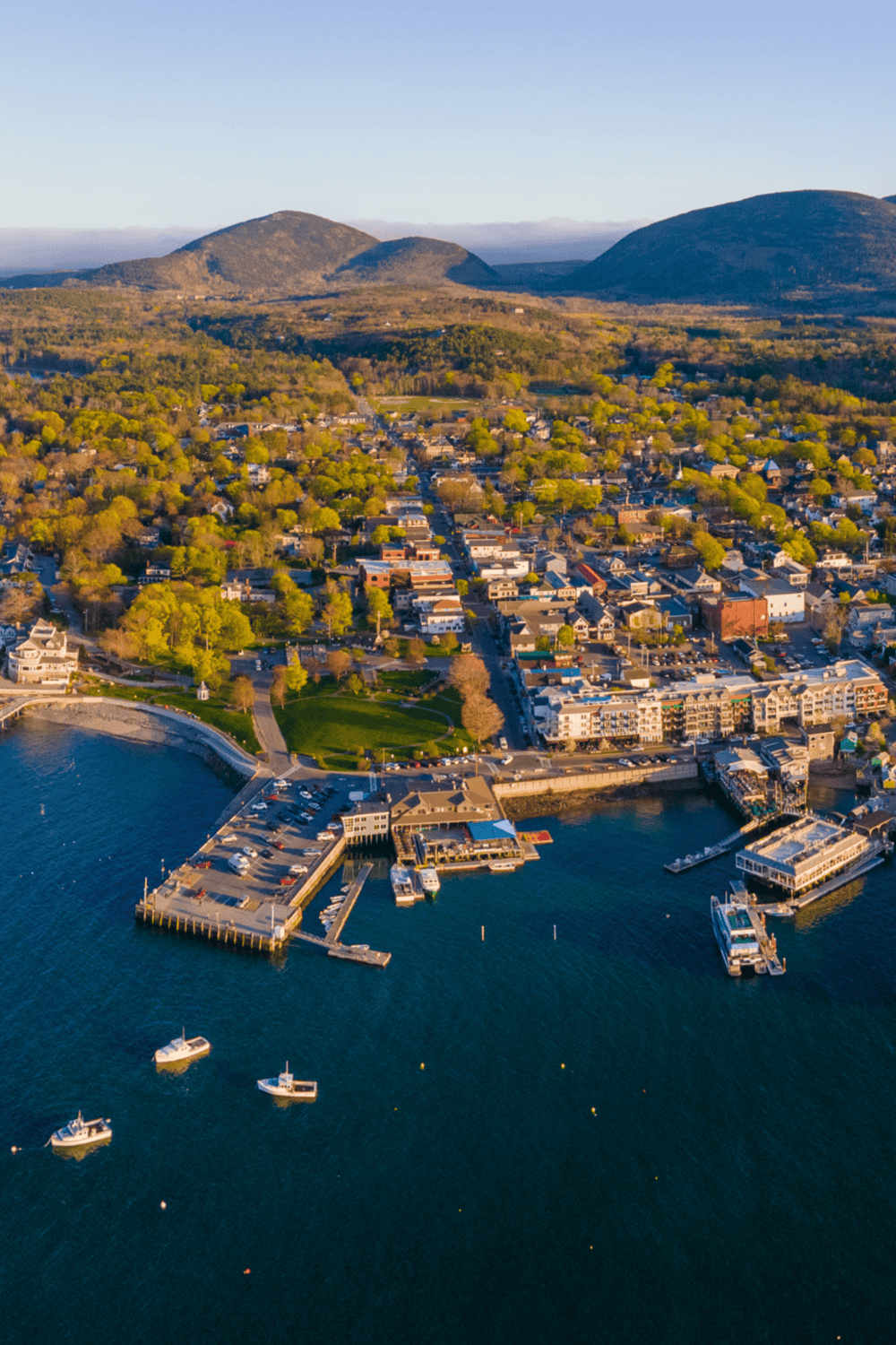 Aerial view of waterfront town with mountains, boats, and lush greenery in the background.
