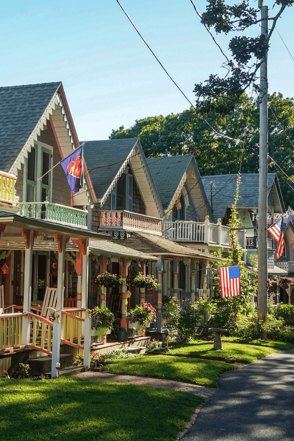Charming colorful Victorian-style houses with American flags on porch, lush greenery, and tree-lined street on a sunny day.