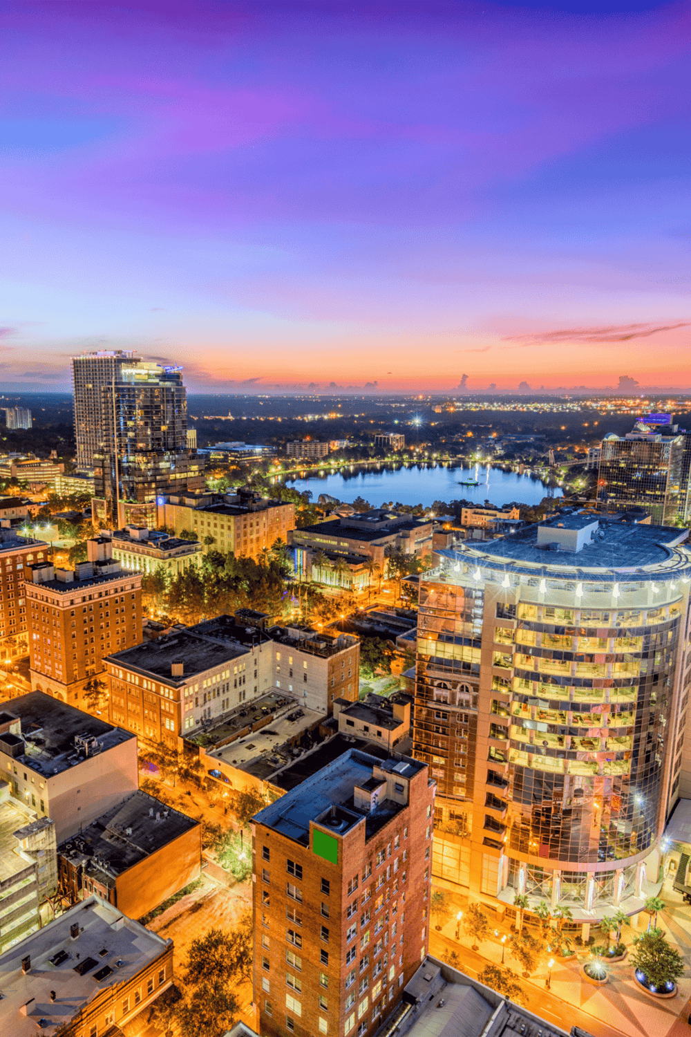 Bright cityscape with high-rise buildings, lake, and colorful sunset sky.