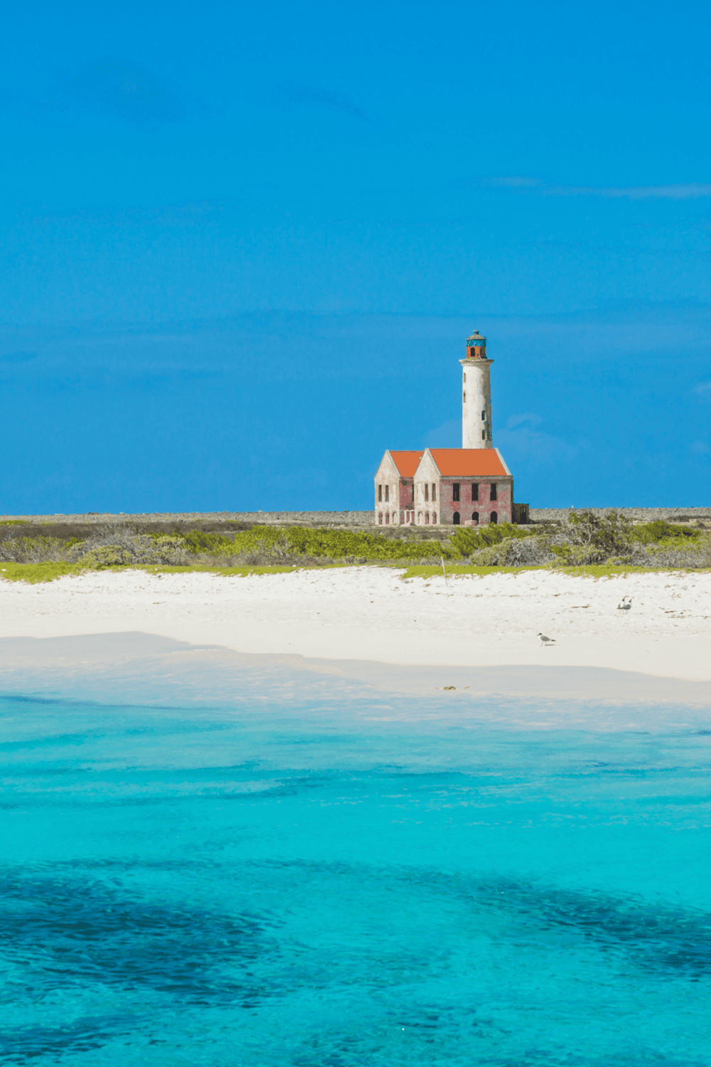 Lighthouse on tropical beach with clear blue water and sandy shore, scenic coastal landscape.