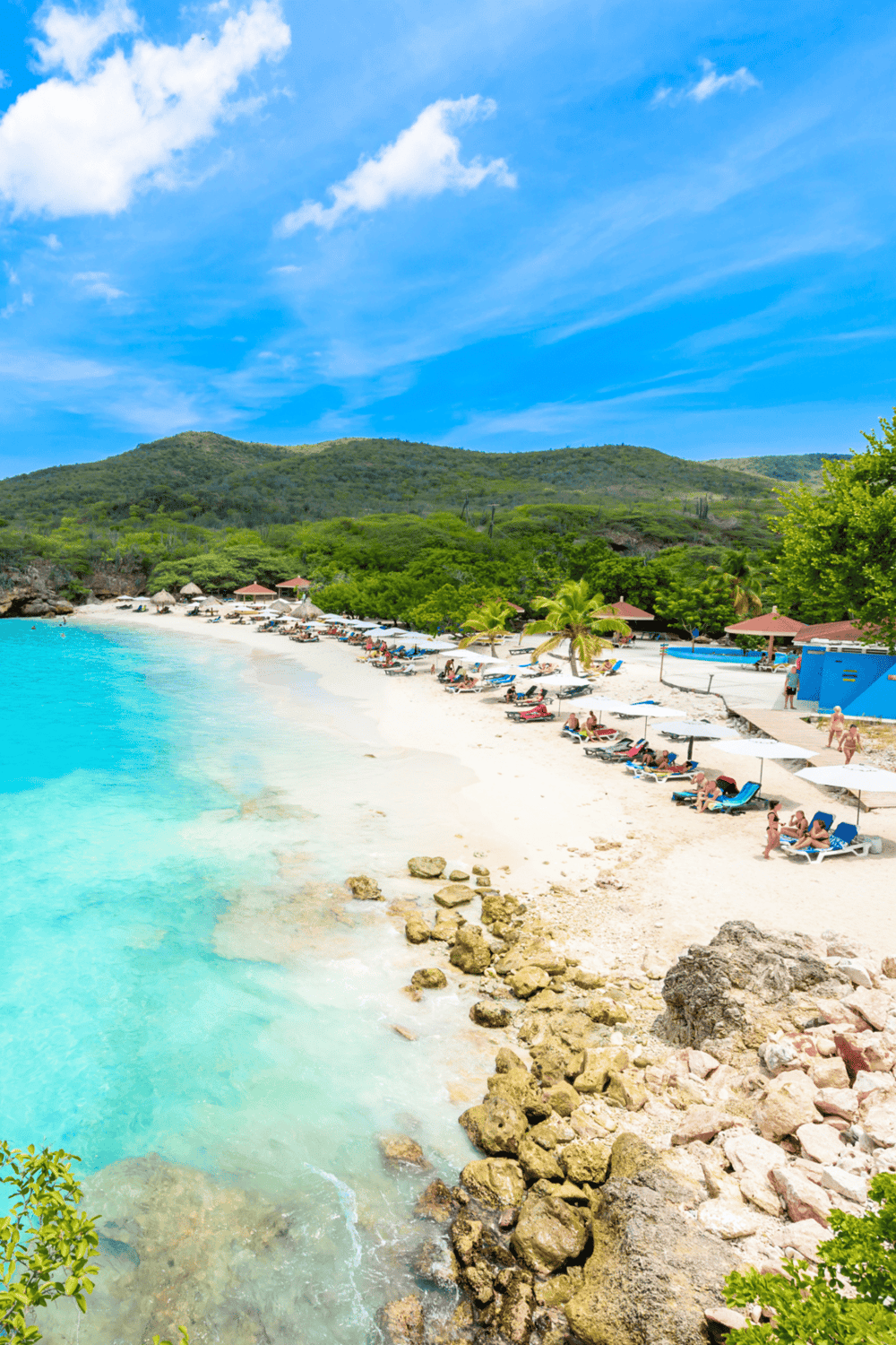 Beach with turquoise waters and sun loungers, lush green hills in the background, perfect for travel guidance.