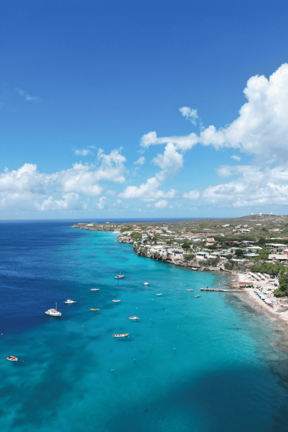 Aerial view of a tropical coastline with turquoise waters, boats, and coastal homes under a blue sky.