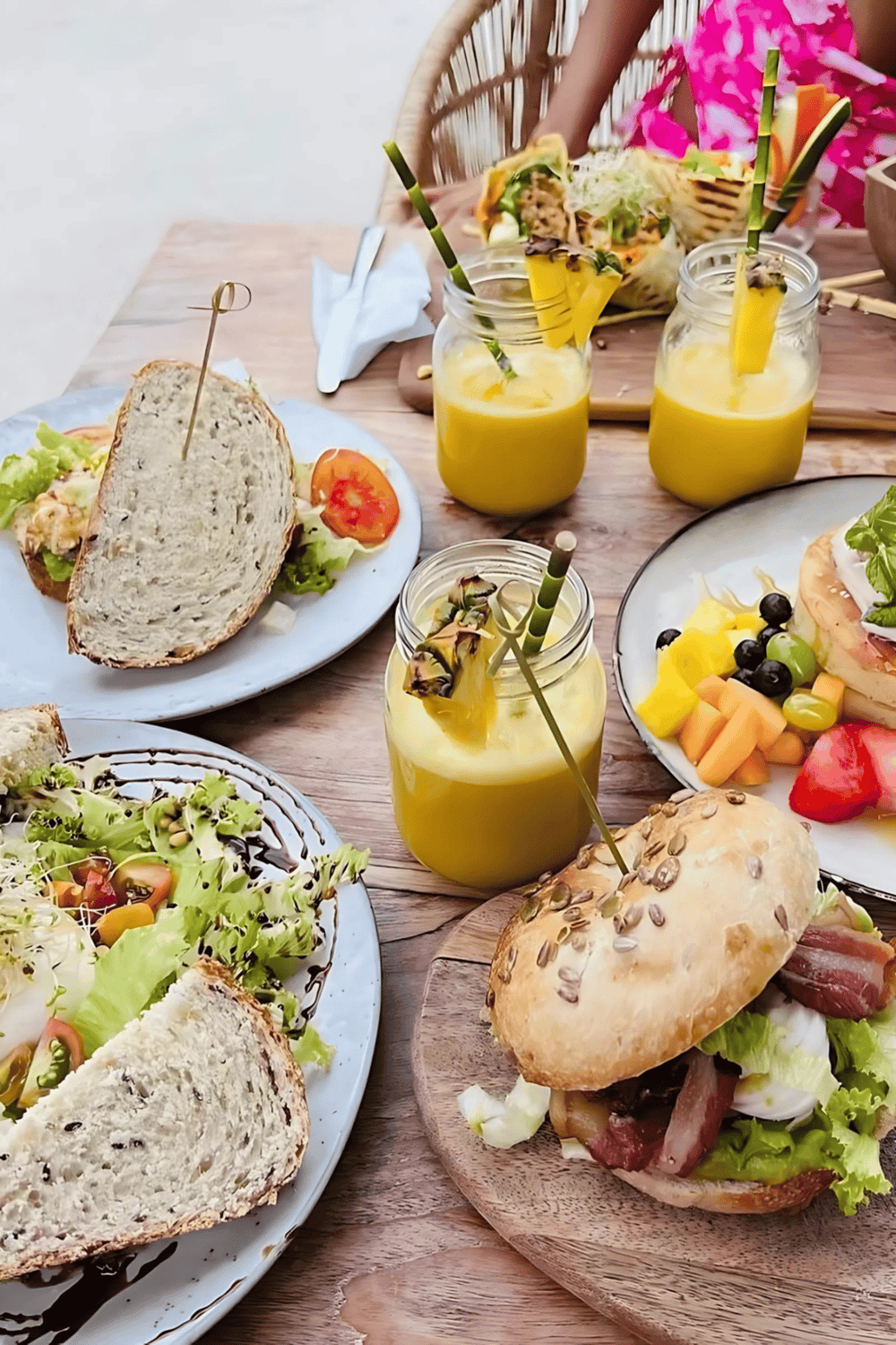 Colorful tropical brunch with smoothies, fruit, salads, and sandwiches on a wooden table.