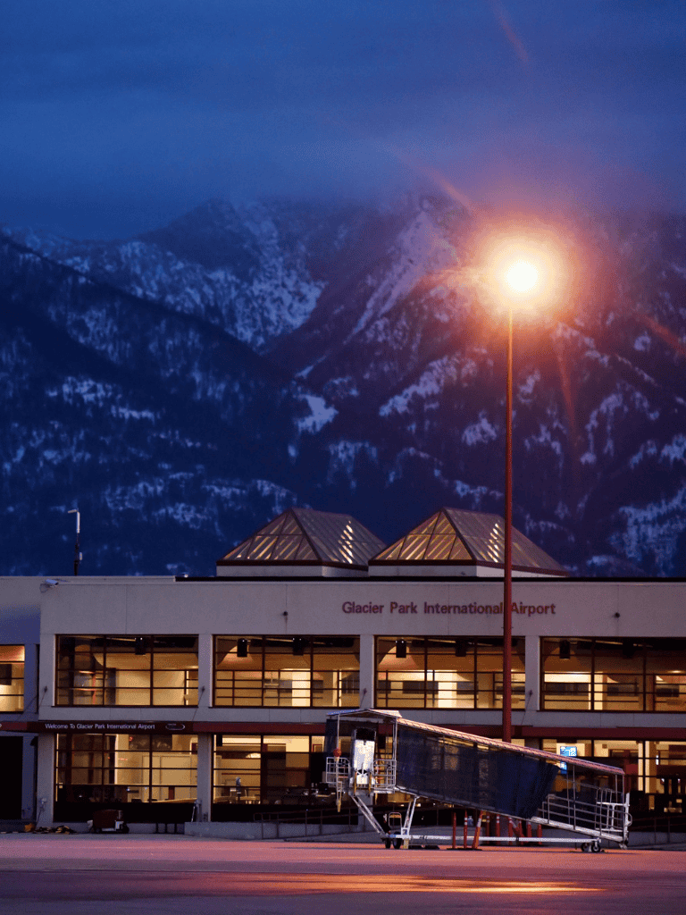 Empty Glacier Park International Airport at dusk with mountain backdrop, airport building, and ramp equipment.
