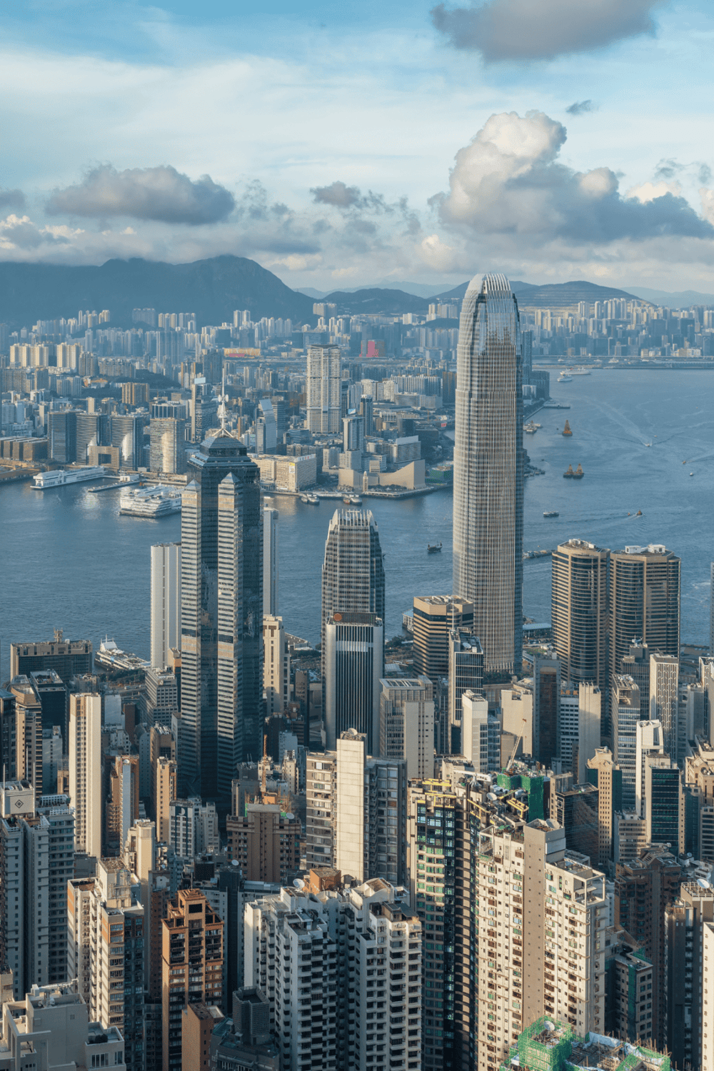 Panoramic view of Hong Kong skyline with skyscrapers, Victoria Harbour, and mountains in the background.