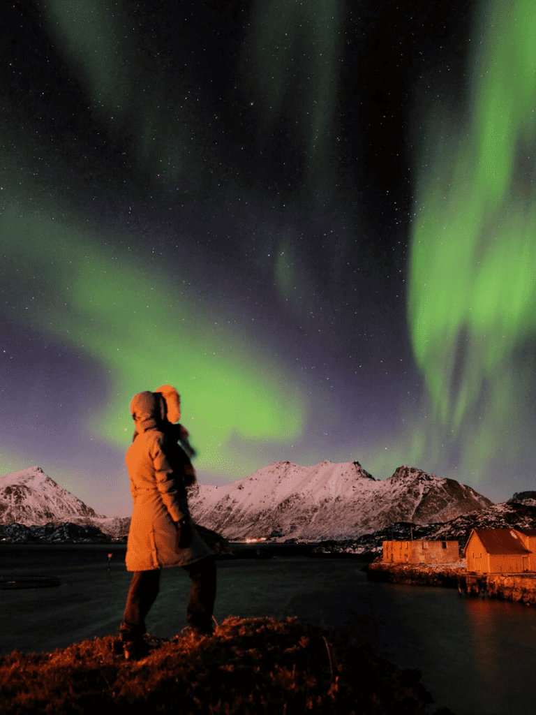 Vivid northern lights over snow-capped mountains with traveler observing in Norway.