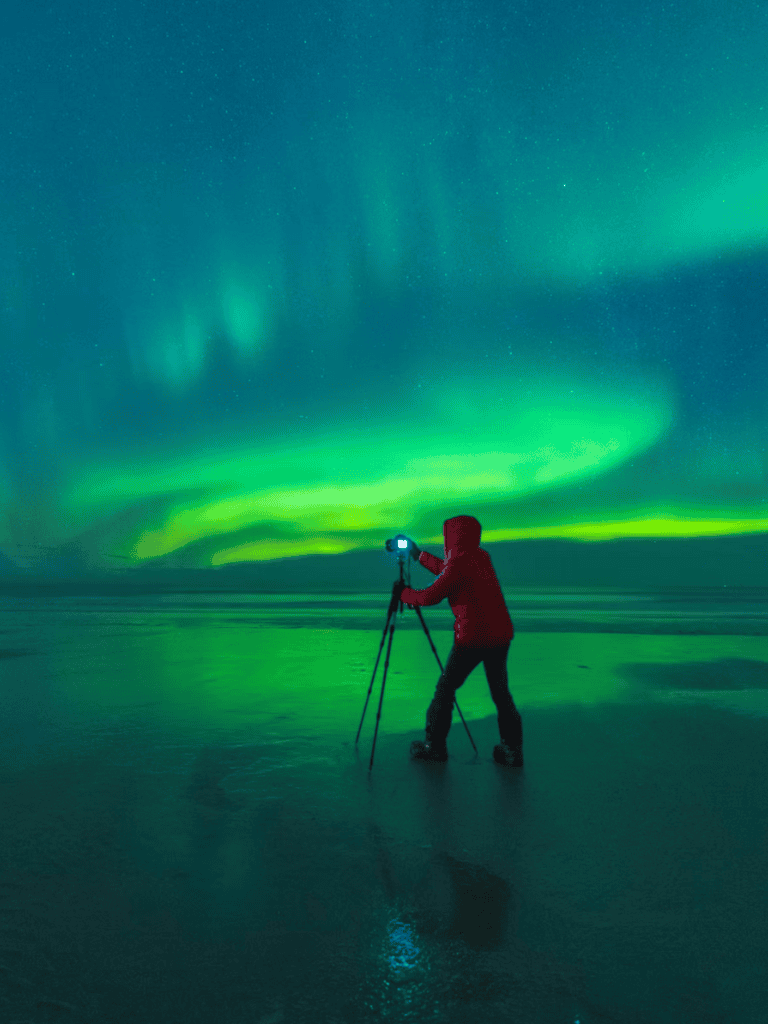 Aurora Borealis over icy landscape with photographer capturing northern lights, Iceland adventure travel, stunning aurora view.