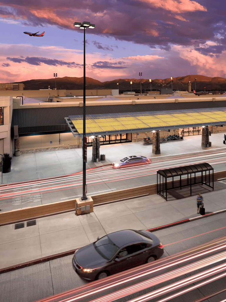 Airplane flying over modern airport terminal at sunset, with light trails from cars and a traveler waiting at the bus stop.