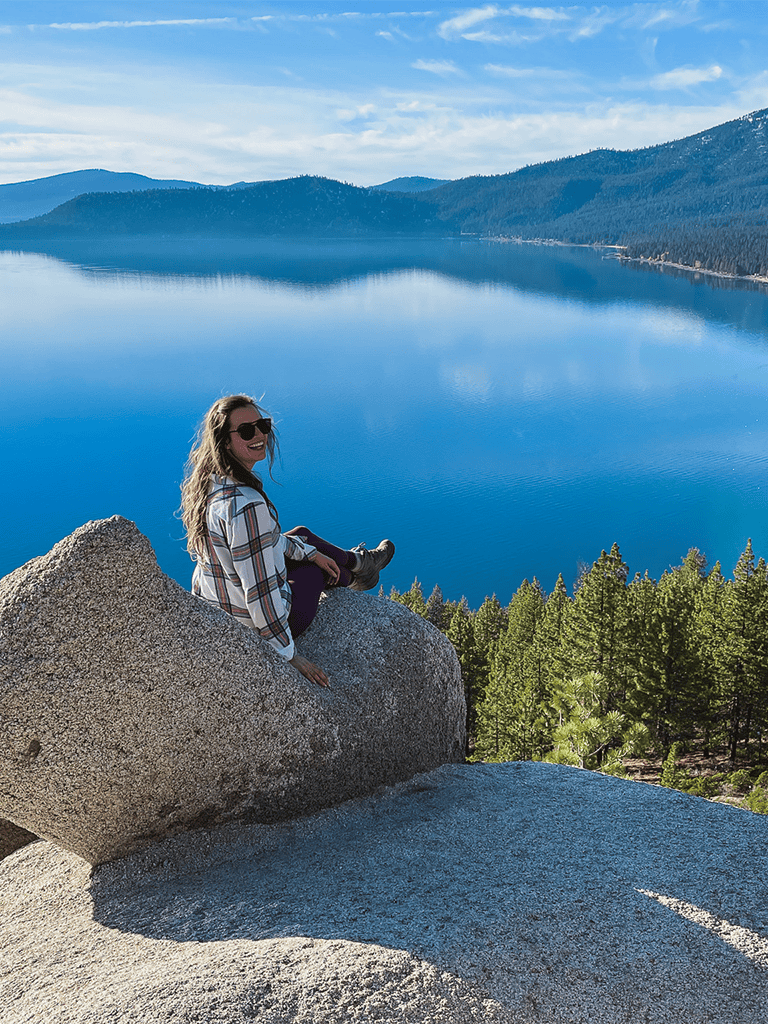 Serene lake scene with a woman sitting on a large rock overlooking the water and mountains.