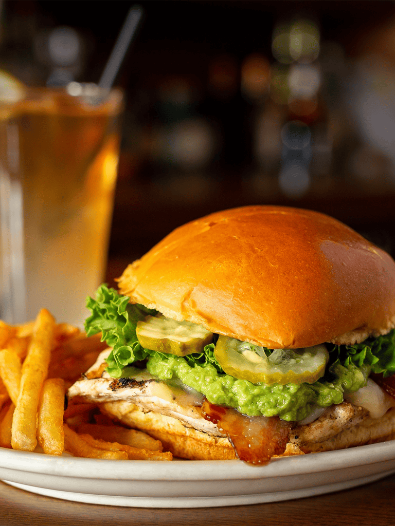 Juicy chicken sandwich with lettuce, pickles, guacamole, and crispy fries on a white plate.