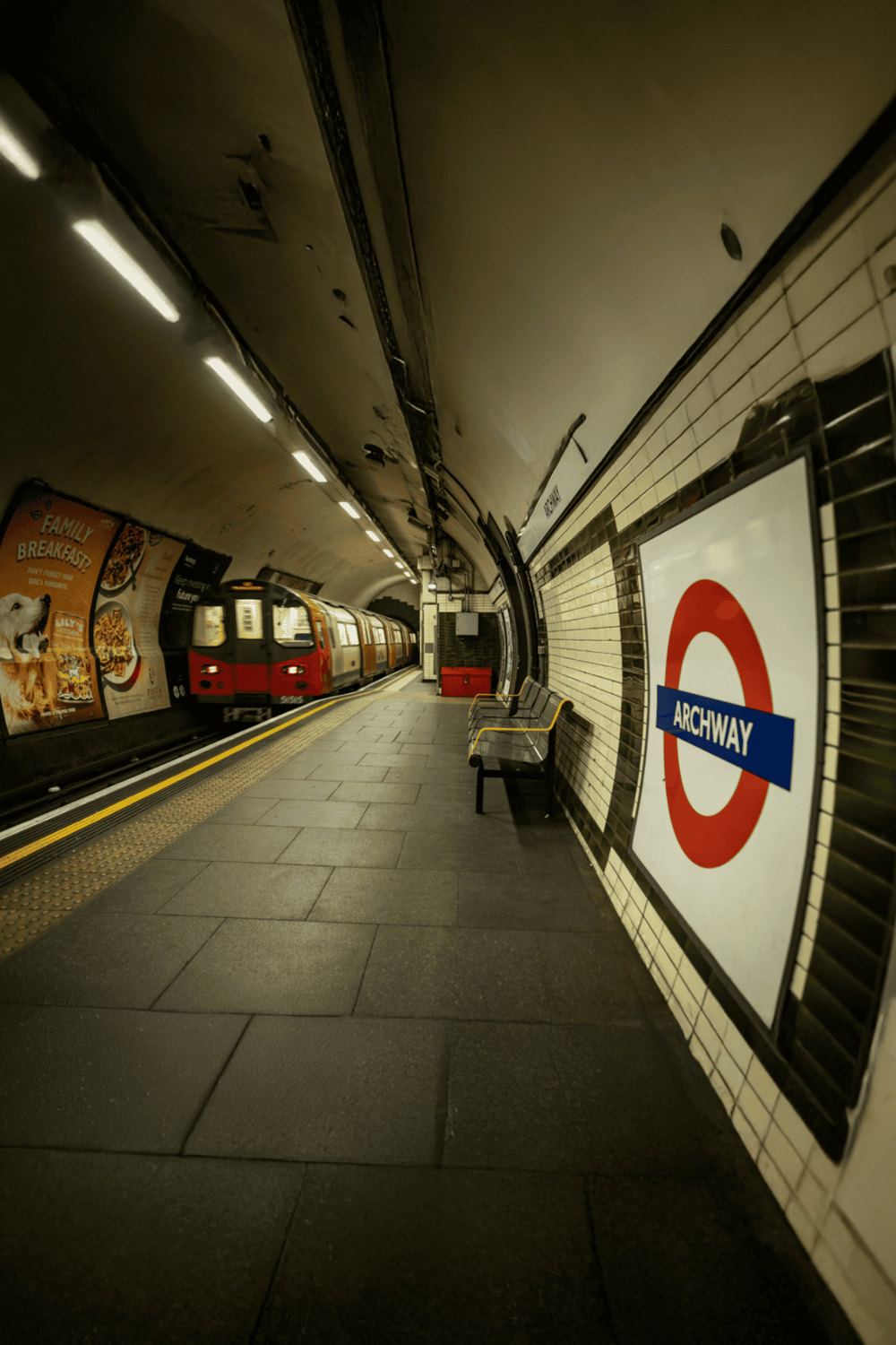 Underground London subway station platform with train arriving, tiled walls, and seating areas.