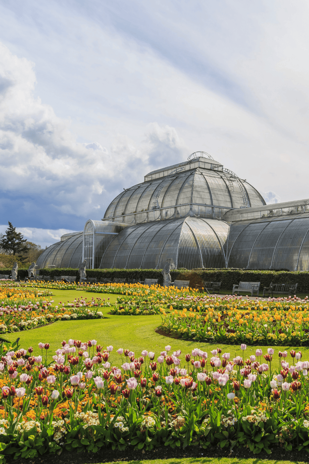 Vibrant flower garden in front of a historic greenhouse or conservatory, lush greenery and colorful blooms.