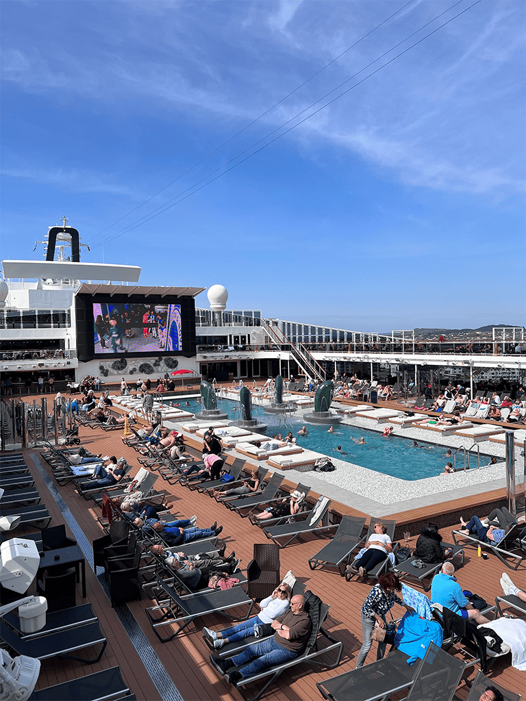 Relaxing pool deck on a cruise ship with loungers and a large outdoor screen under a clear blue sky.