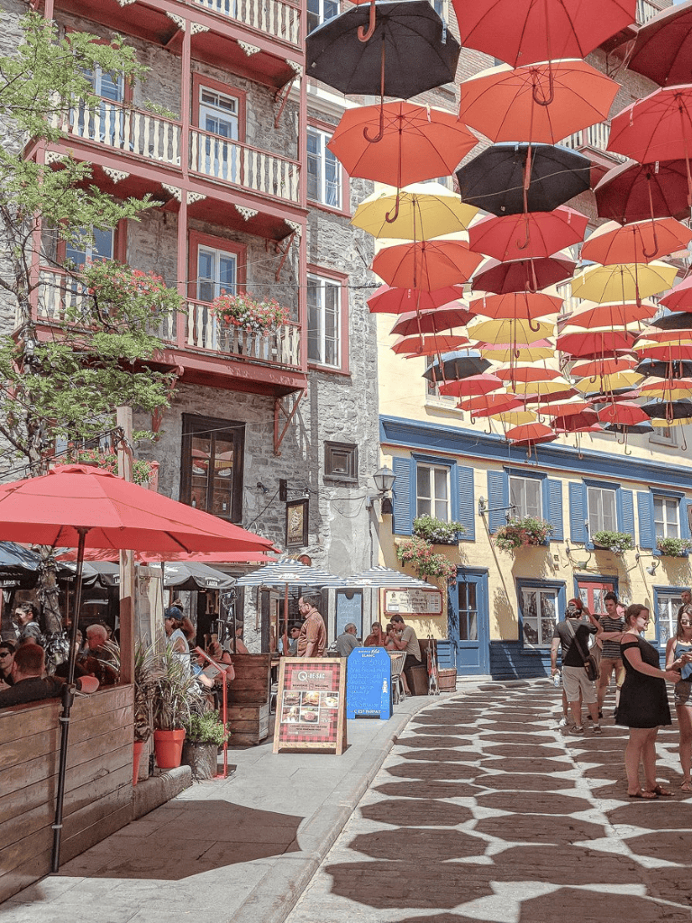 Colorful umbrellas overhead in a lively outdoor cafe in a charming European street scene.