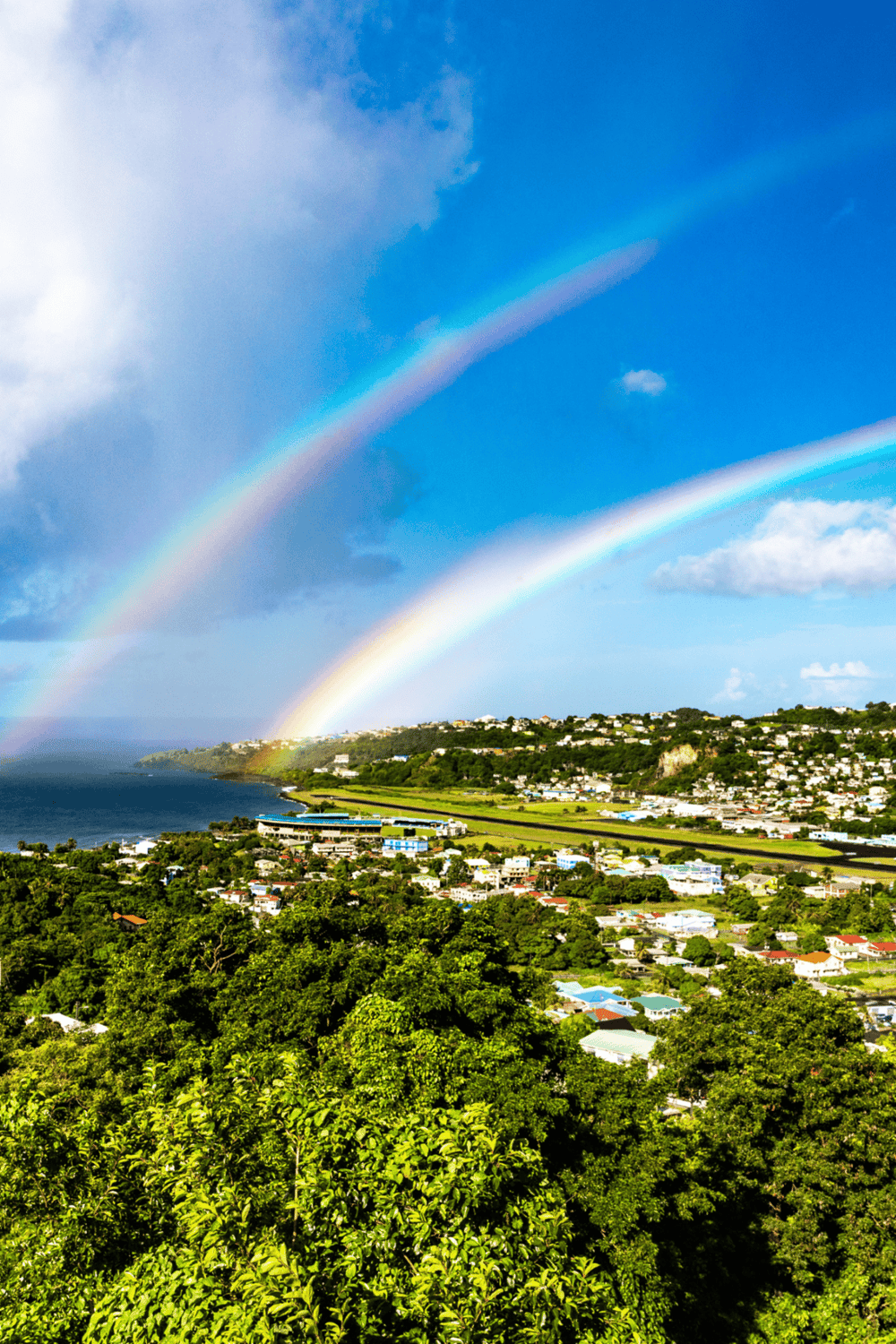 Vivid double rainbow over lush green island town, scenic ocean view in the Caribbean.