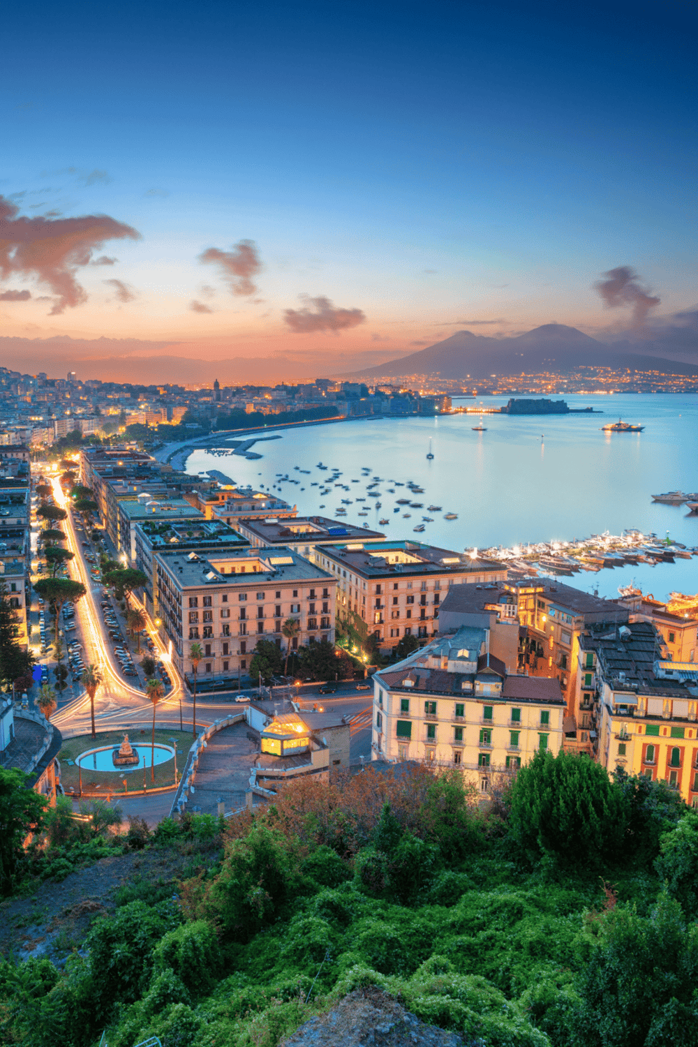 Spectacular view of Naples by the bay at sunset, showcasing vibrant city lights and Mount Vesuvius in the background.