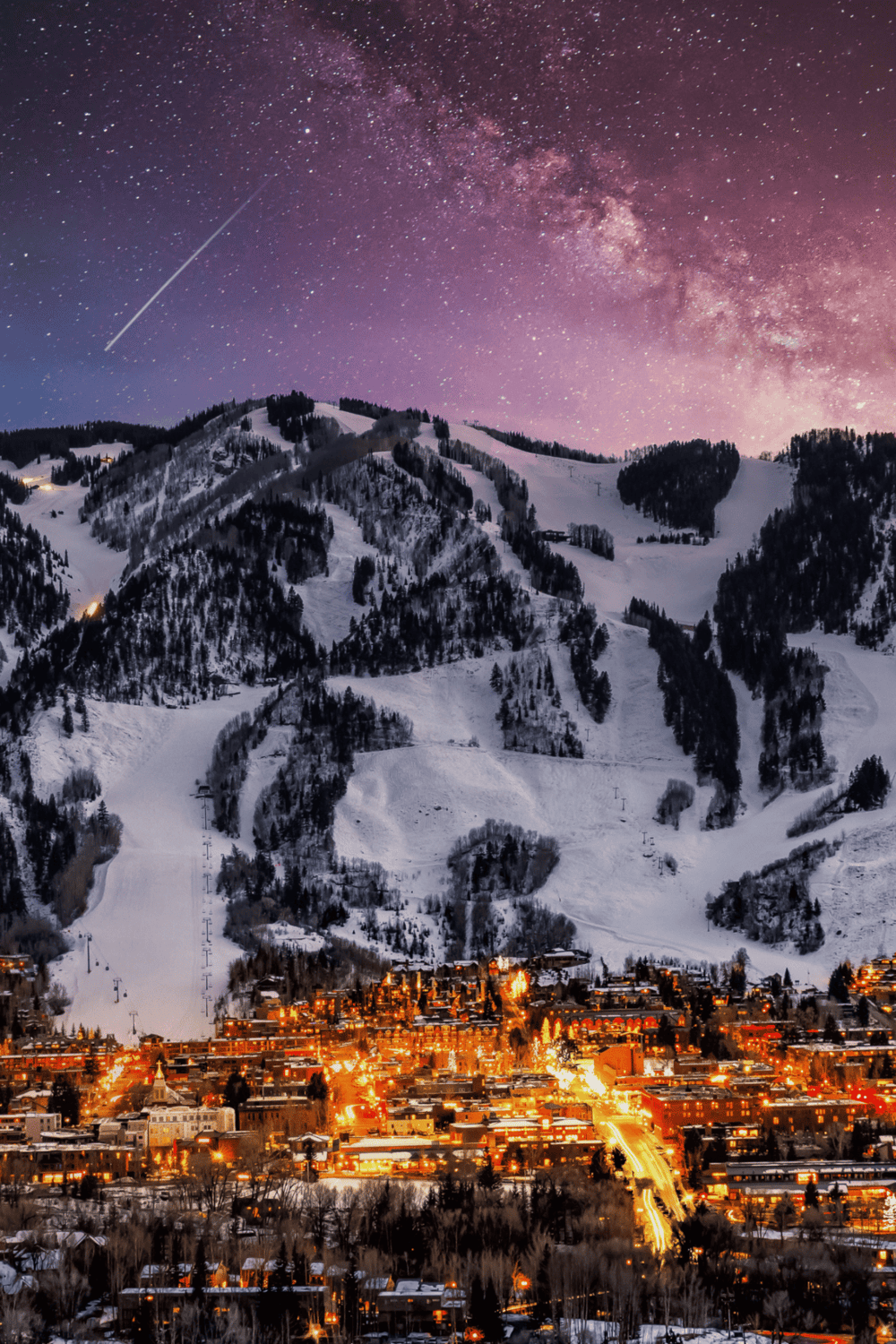 Stargazing over snowy mountain town with city lights under a starry night sky.
