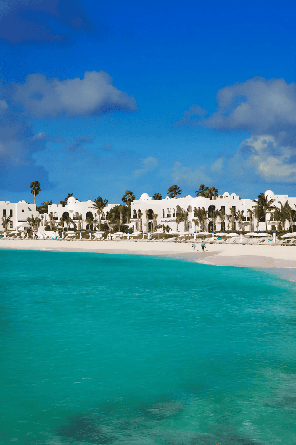 Relaxing beach resort with white buildings, palm trees, and turquoise waters under a bright blue sky.