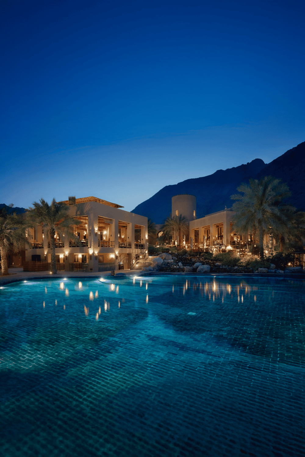 Luxurious desert resort with illuminated pool and mountain backdrop at dusk.
