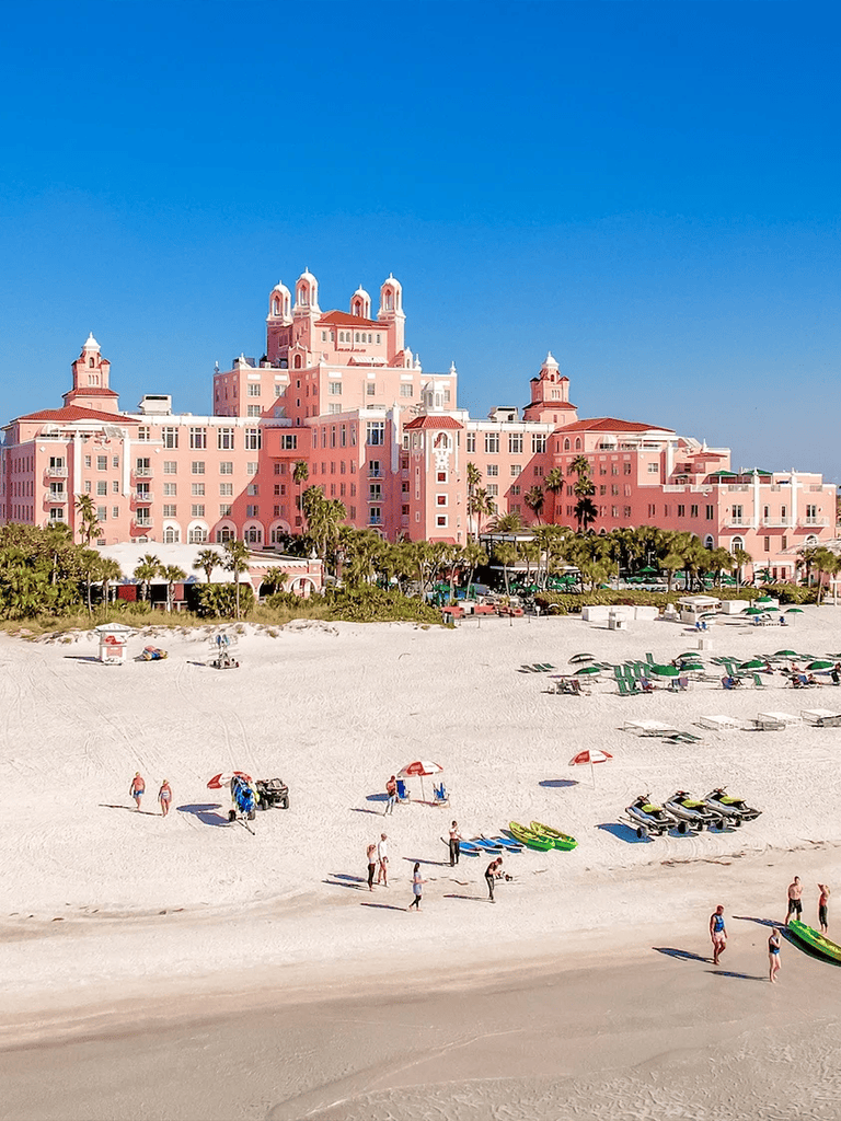 Pink hotel on beach with paddleboards and umbrellas under bright blue sky.