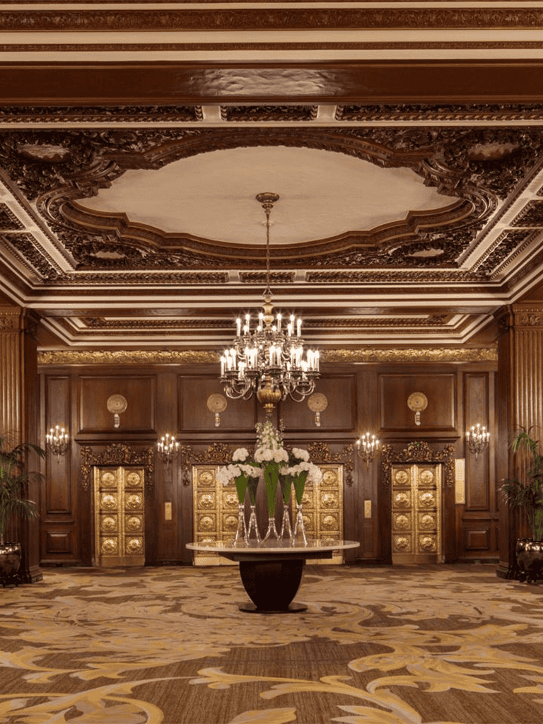 Luxurious hotel lobby with ornate woodwork, chandelier, and elegant floral centerpiece.