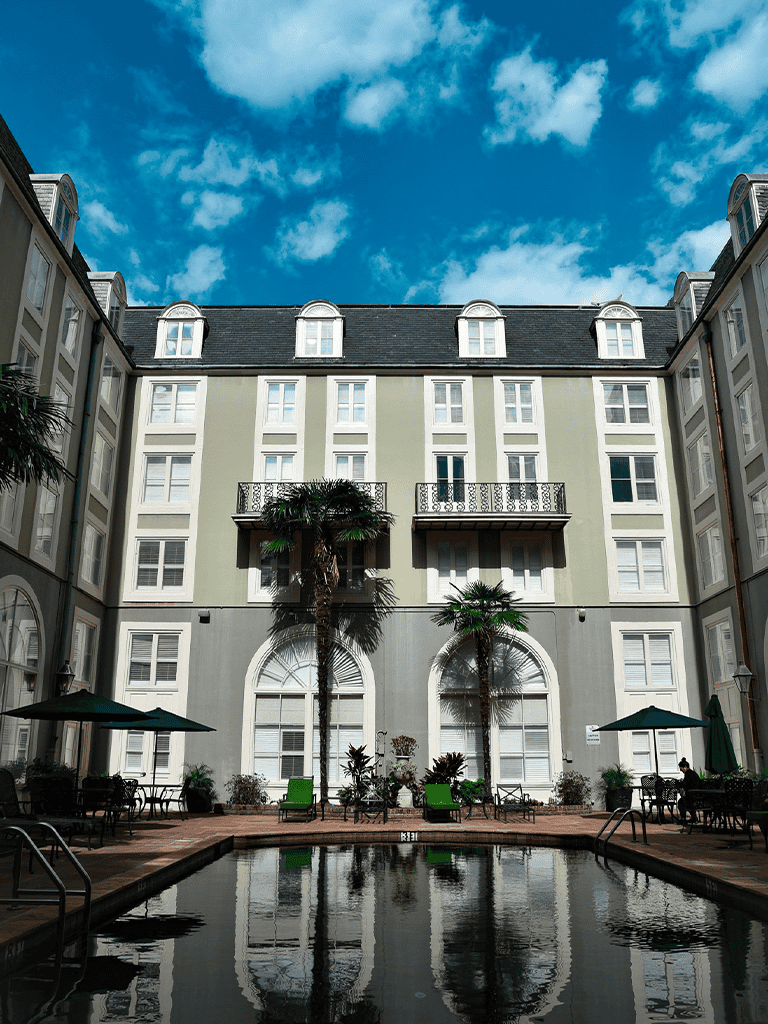 Palm trees and pool inside hotel courtyard with Mediterranean architecture, blue sky, and cloud background.