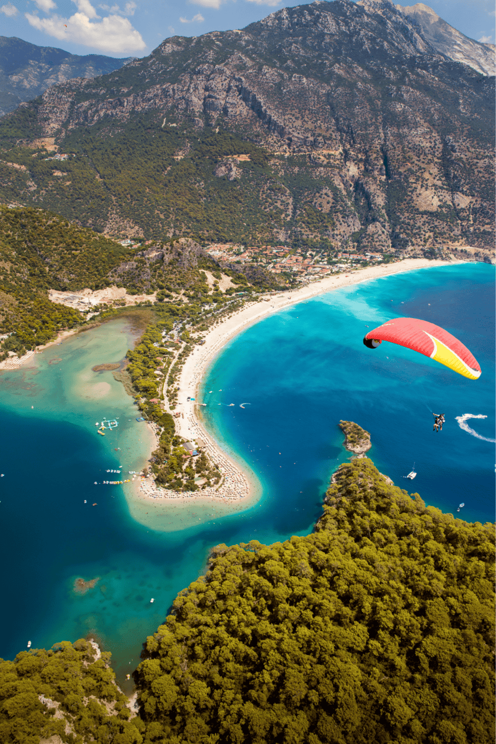 Vibrant coastal scenery with paragliding over a crescent-shaped beach and turquoise water, surrounded by lush greenery and mountainous terrain.