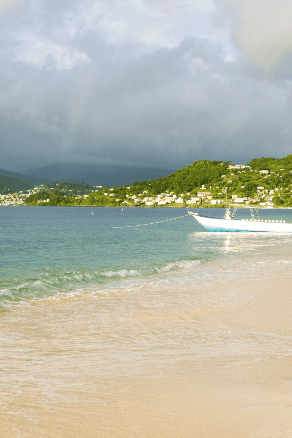 Sunlit tropical beach with calm waters and boat near lush hillside town, perfect for vacation travel.