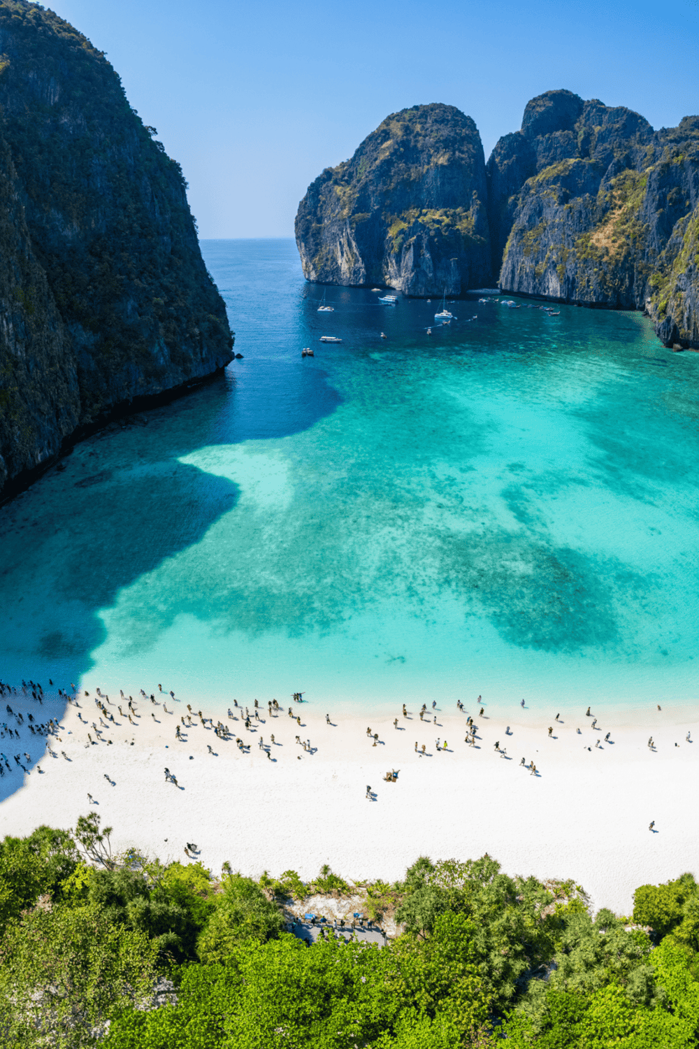 Stunning tropical beach with turquoise waters, towering limestone cliffs, and boats at anchor.