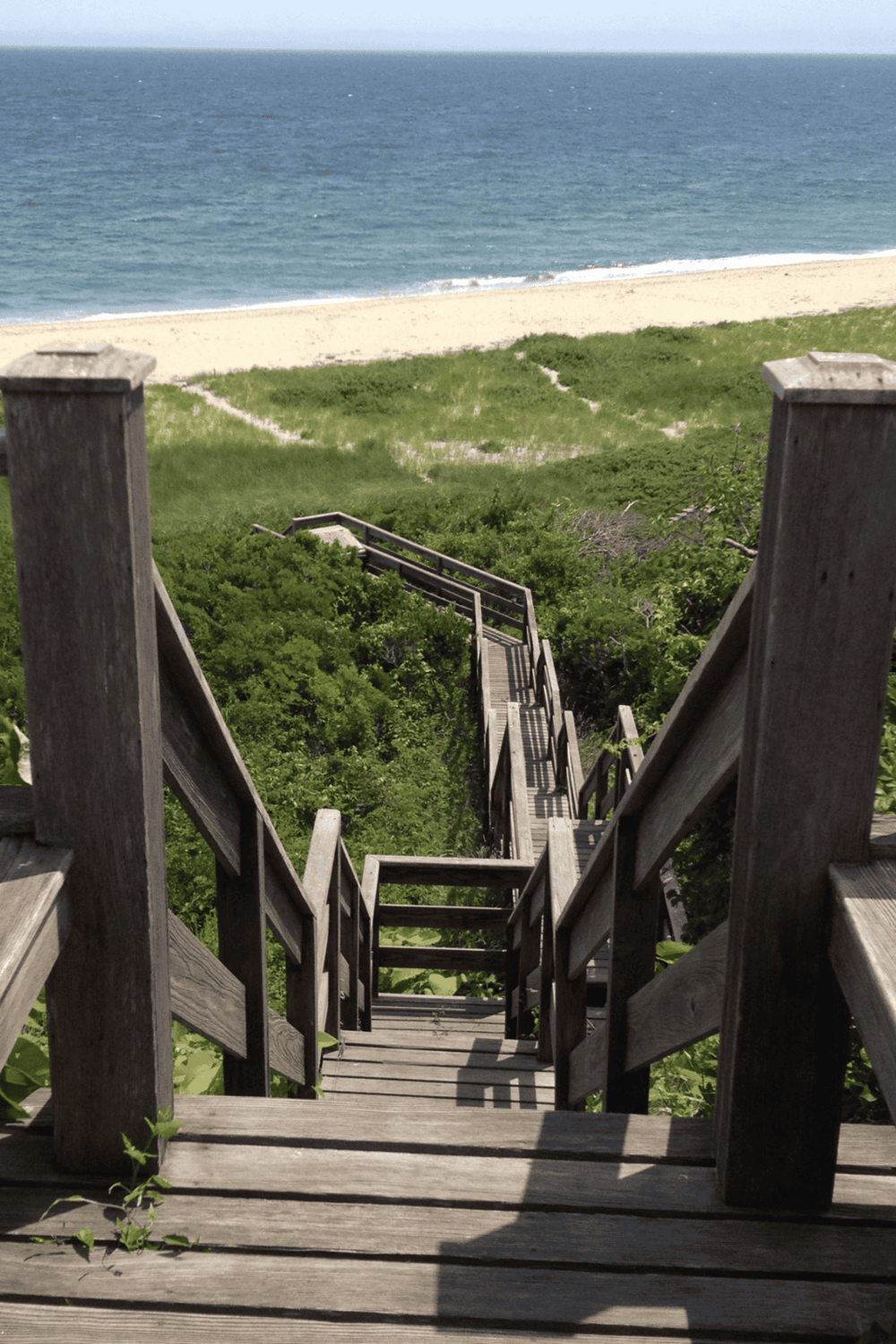 Steep wooden staircase leading down to a sandy beach with ocean view and lush green surroundings.