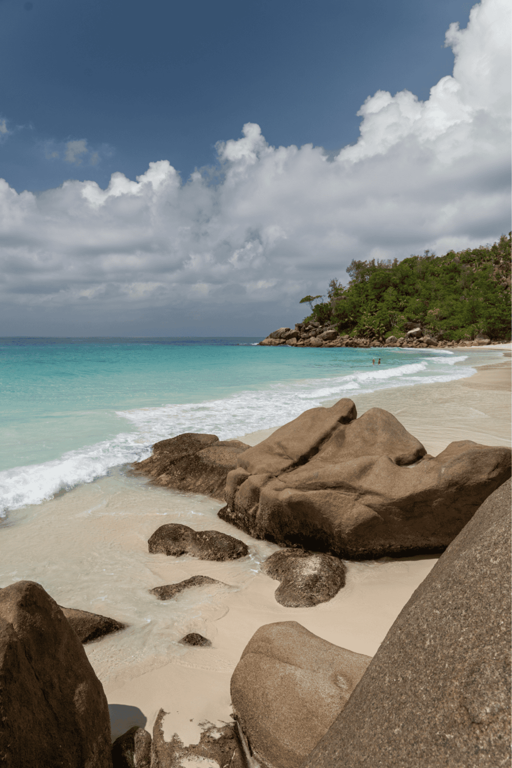 Cloudy beach scene with rocks, turquoise water, and green hillside, perfect for vacation destinations.