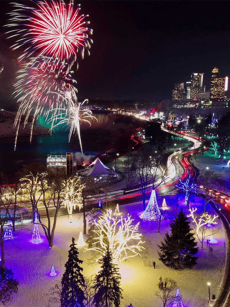 Fireworks display over a city skyline with festive holiday lights and illuminated trees in winter.