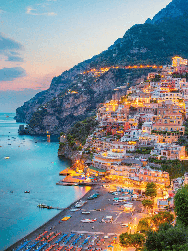 Colorful seaside village on cliffside with boats and beach at sunset.