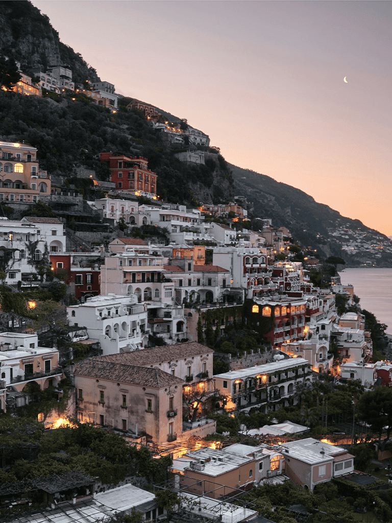 Colorful hillside town at sunset in Amalfi Coast, Italy, with illuminated buildings and scenic coastal views.
