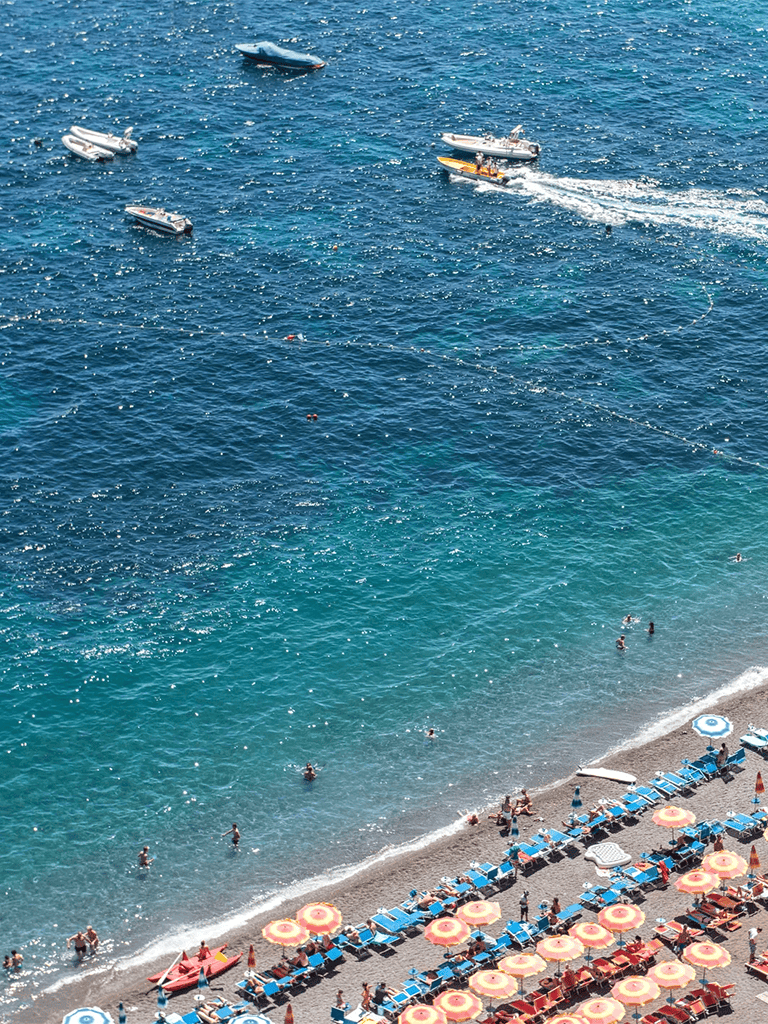 Boats in blue ocean water with beach and umbrellas.