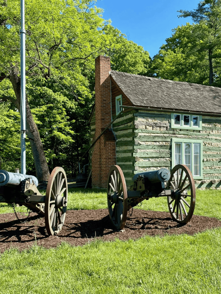 Cannons in front of historic log cabin surrounded by greenery and bright blue sky.