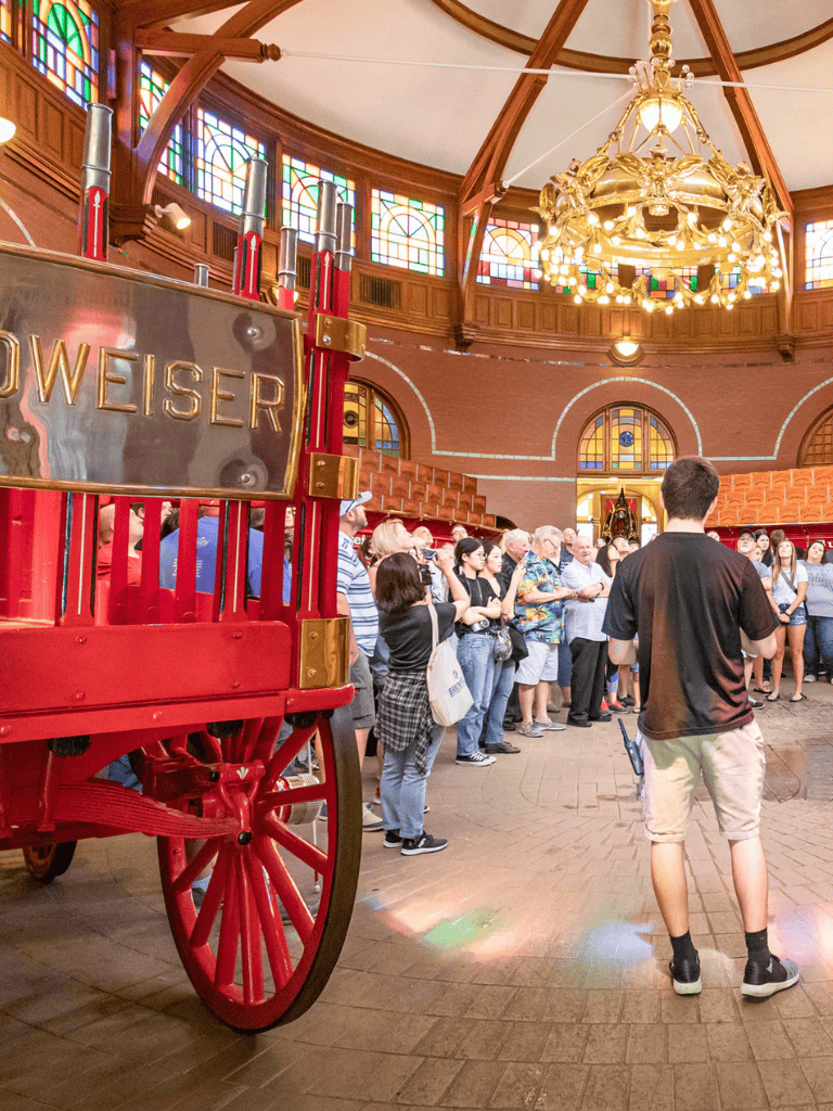 Colorful indoor event space with a large group of people and a vintage Budweiser cart in the foreground.