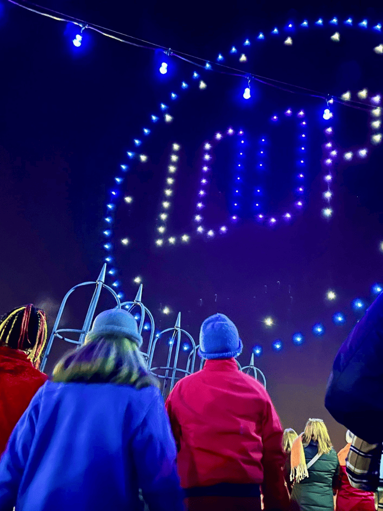 Colorful holiday lights display reading "JOY" at night. People gather under festive illuminations for joyful celebrations.