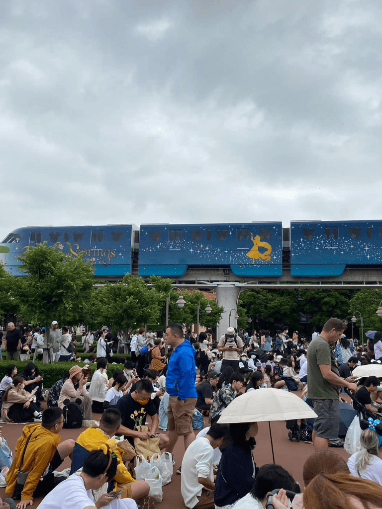 - Excited crowd waiting at Disney park with Mickey Mouse monorail overhead, cloudy sky.