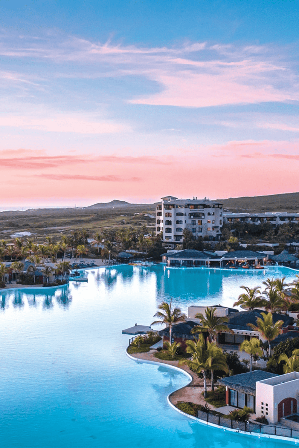 Bright sunrise over luxurious resort pool with palm trees and hillside in background.