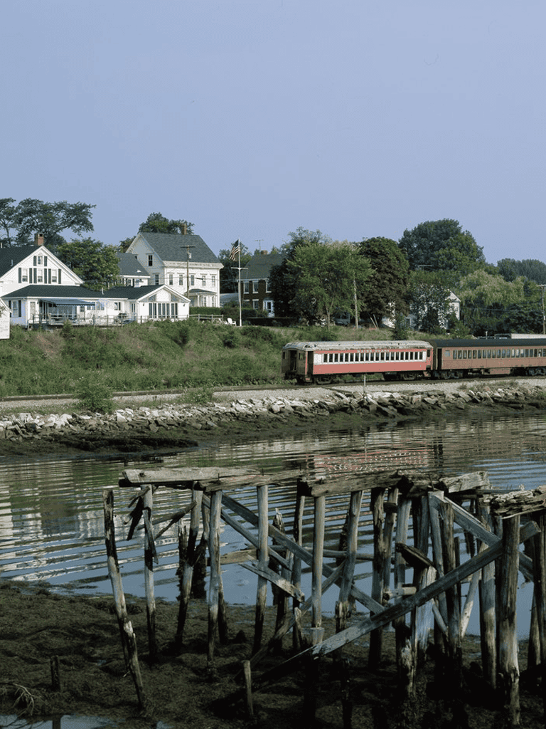 Old train passing by waterfront with houses and a wooden pier in the foreground, scenic rural landscape.