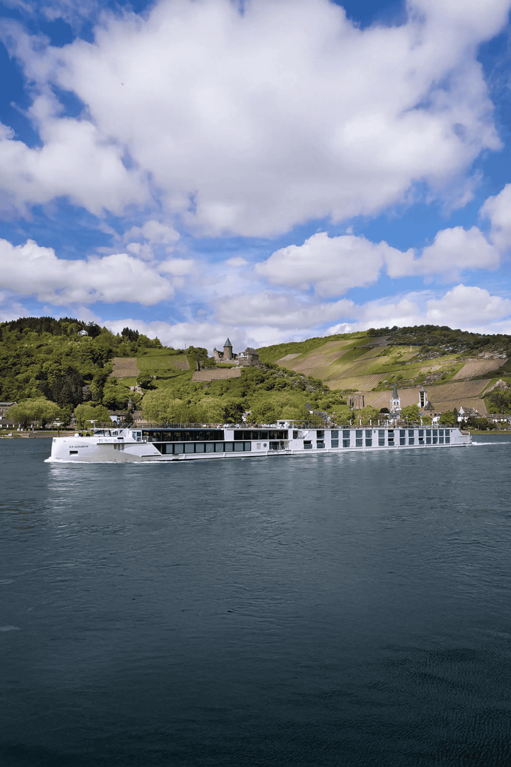 Cruise ship sailing on a river with scenic hillside vineyards and historic buildings in the background.