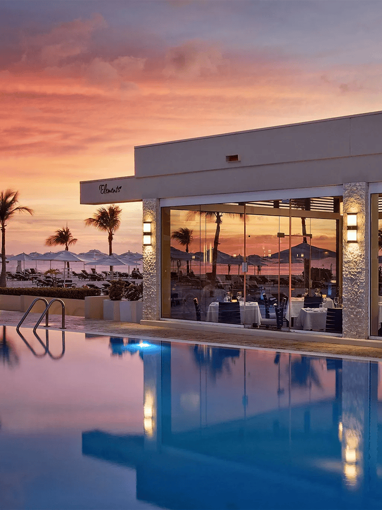 Luxury beachfront hotel poolside dining at sunset, palm trees, 5-star resort atmosphere, ocean view, Elements restaurant.