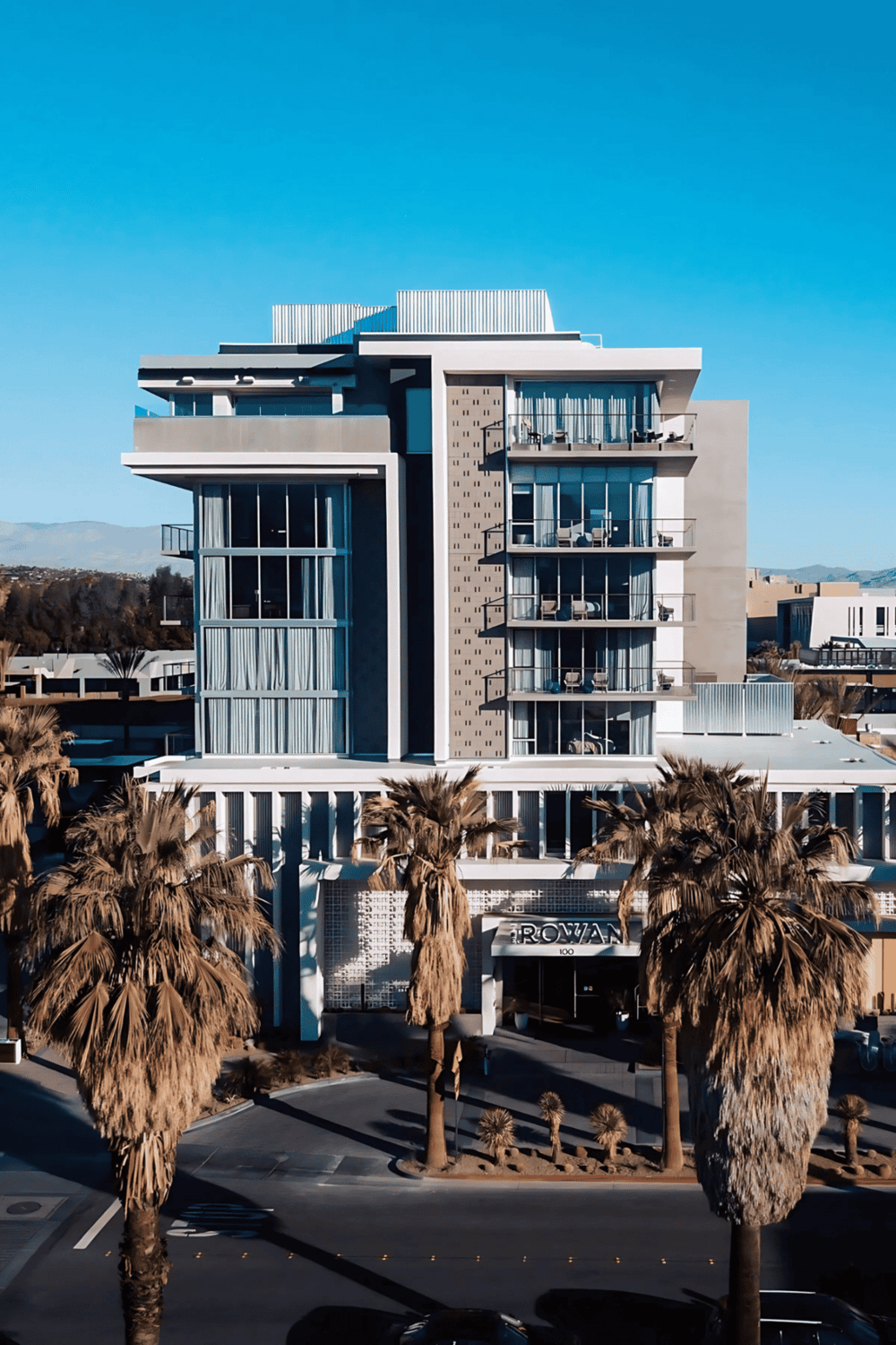 Modern luxury hotel building with palm trees in front, scenic mountains, and clear blue sky.