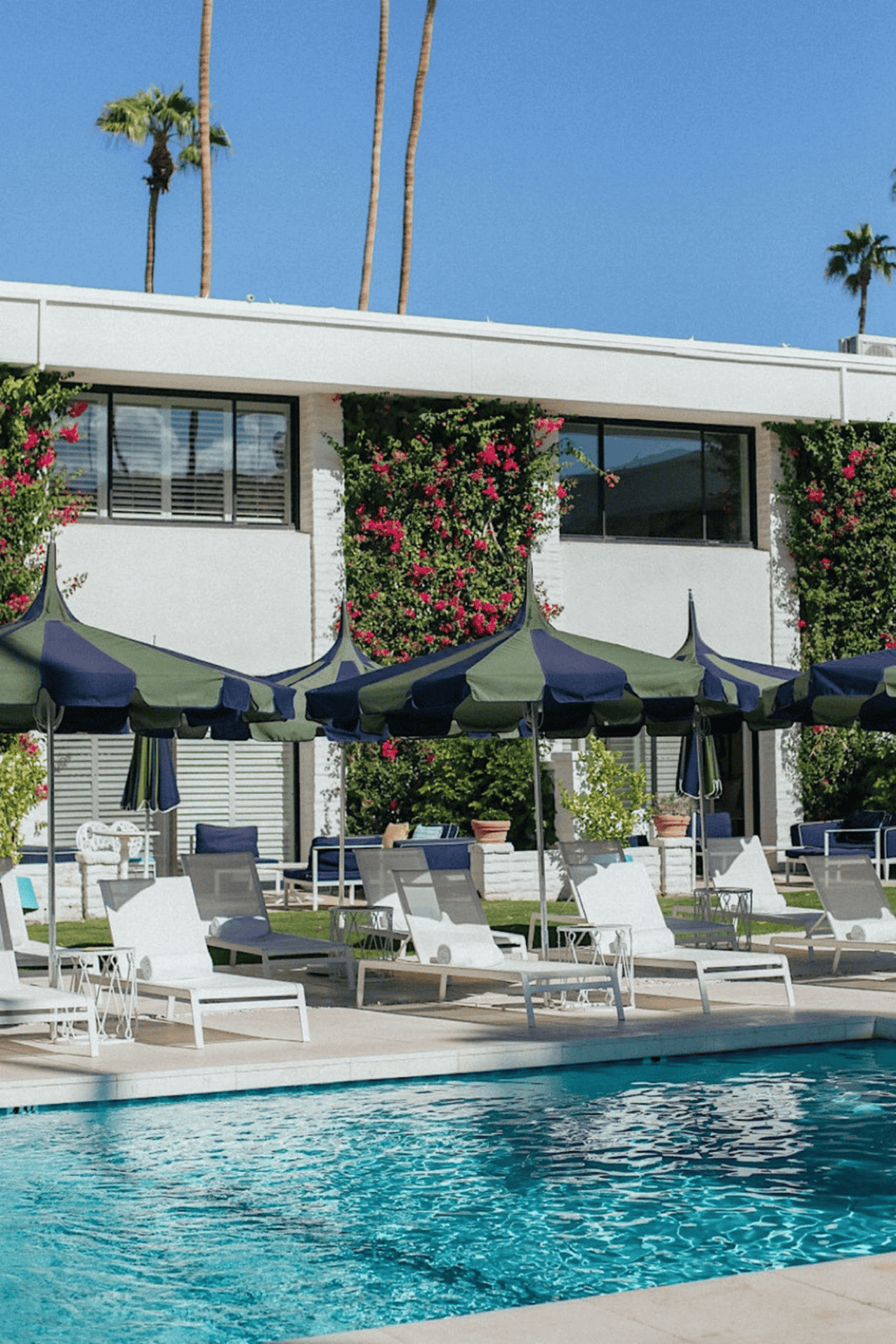 Sunlit hotel pool area with lounge chairs, umbrellas, and lush greenery under a clear blue sky.
