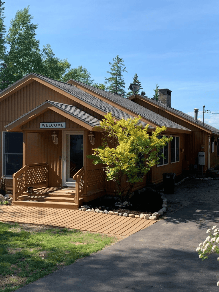 Cozy wooden house with "Welcome" sign, surrounded by greenery and a sunny pathway.