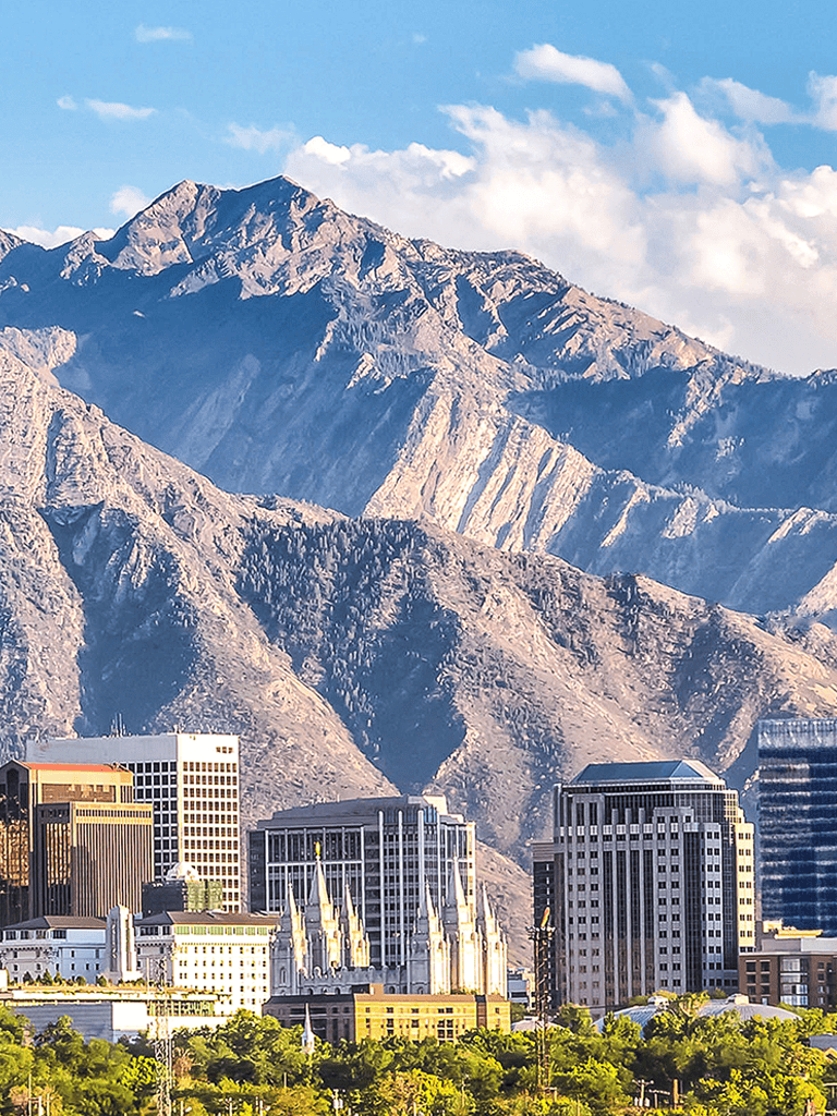 Vibrant mountain city skyline with modern buildings and historic architecture, Utah mountains in background, sunny day.