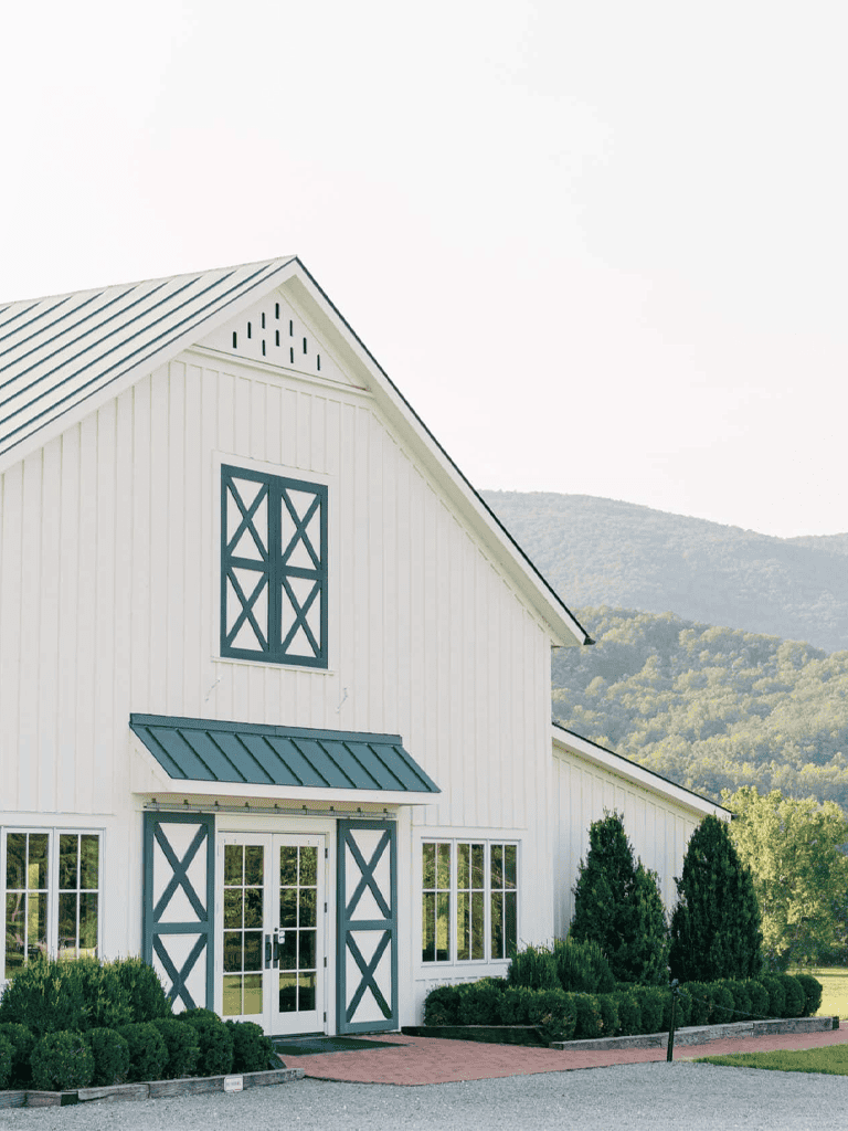 Ceramic barn with modern farmhouse style, white exterior, blue accents, surrounded by green landscape and mountains.