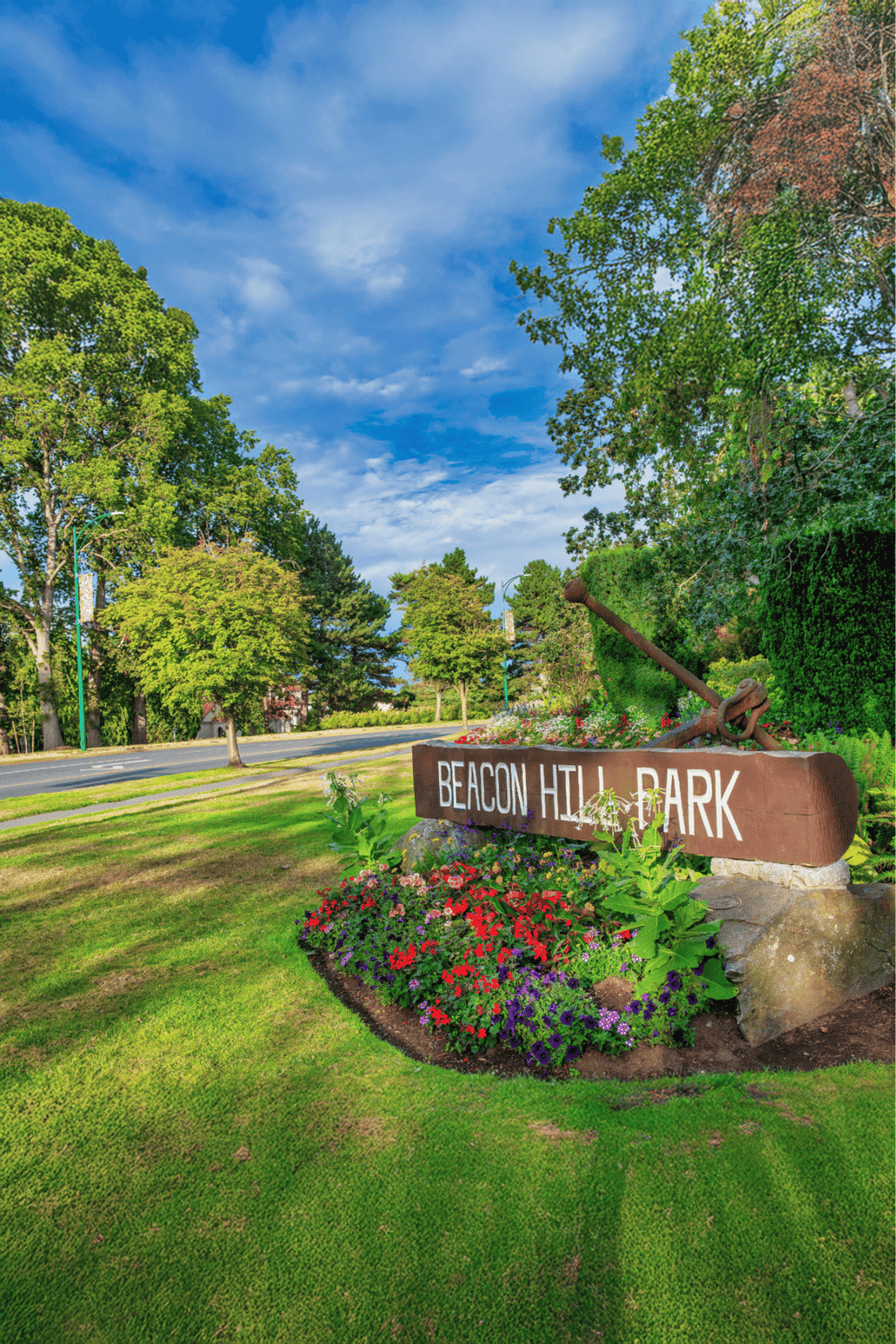 Lush green park with vibrant flowers, Beacon Hill Park sign, and scenic trees under a clear blue sky.